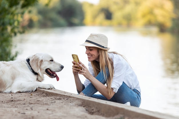 Foto de una joven fotografía aficionada que toma una foto de su perro sentado cerca del río en el parque.