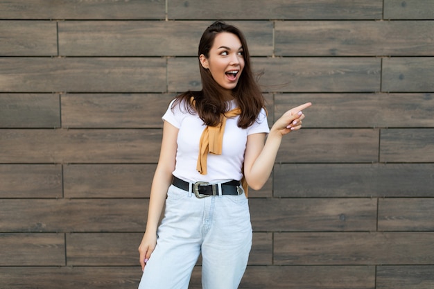 Foto de joven hermosa morena sorprendida positiva emocional vistiendo ropa casual de pie en la calle cerca de la pared y apuntando al espacio vacío. Concepto de estilo de vida.