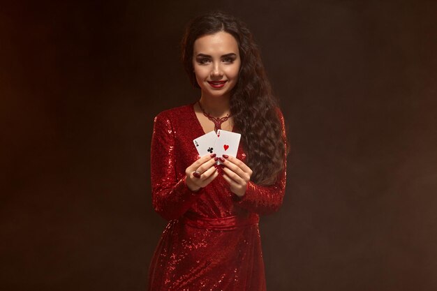 Foto de joven hermosa mujer morena caucásica en vestido de noche rojo brillante, sosteniendo cartas frente a ella con ambas manos, muestra un par de ases. Fondo marrón, casino, industria del juego