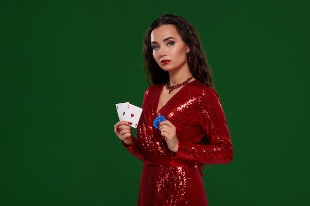 Foto de joven hermosa mujer morena caucásica en vestido de noche rojo brillante tiene sus cartas, un par de ases y algunas fichas en sus manos. Fondo verde, chromakey, casino, industria del juego