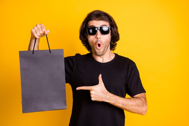 La foto del joven indica el dedo directo bolso del centro comercial expresión de asombro boca abierta camiseta gafas de sol aislado fondo de color amarillo