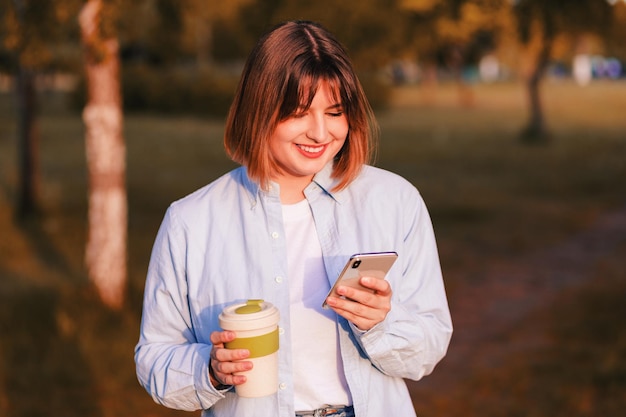 Foto de una joven linda y soñadora que usa una camisa azul sonriendo caminando disfrutando de una sabrosa taza de té y café ecológico