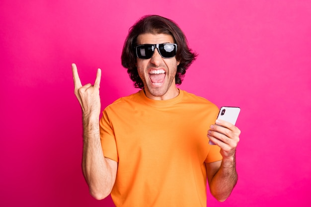 Foto de joven loco mantenga el teléfono con la boca abierta mostrar cuernos usar camiseta naranja gafas de sol fondo de color rosa aislado