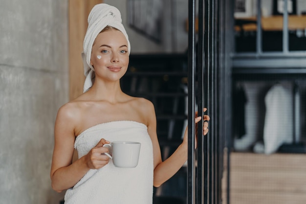 Foto de una joven modelo satisfecha que usa crema en la cara queda satisfecha después de tomar un baño envuelto en una toalla suave blanca bebe café o té posa sobre el interior de la casa Tratamiento antienvejecimiento