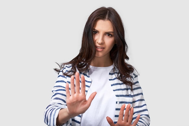 Una foto de una joven morena haciendo un gesto de parada con las palmas de las manos Retrato de una mujer caucásica con una camisa a rayas y una camiseta blanca haciendo un gesto de rechazo posando sobre fondo blanco Negativo