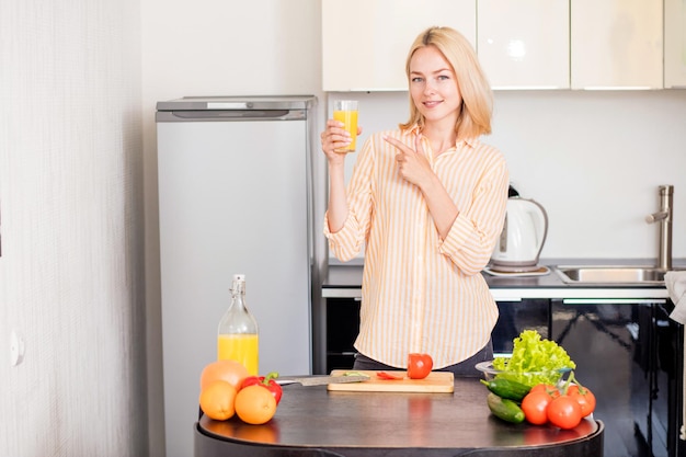 Foto de joven mujer caucásica en camisa a rayas bebiendo jugo recién exprimido mientras se cocina el desayuno. Chica rubia disfrutando de una sabrosa bebida en la cocina de su casa mirando a la cámara con una linda sonrisa.