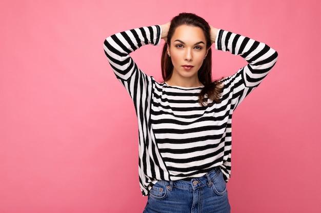 Foto de joven mujer morena hermosa seria segura de sí misma con emociones sinceras vistiendo suéter de rayas casual aislado sobre fondo rosa con espacio de copia.