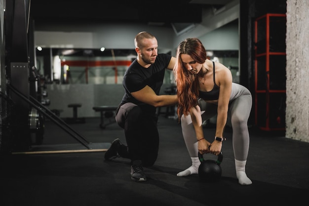 Una foto de una joven musculosa haciendo ejercicio con un entrenador personal en el gimnasio. Ella está haciendo ejercicios con pesas rusas.