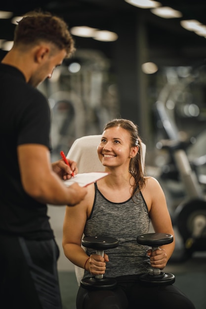 Una foto de una joven musculosa con ropa deportiva trabajando con un entrenador personal en la máquina del gimnasio. Ella está bombeando su músculo con pesas.