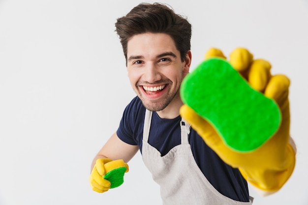 Foto de joven optimista con guantes de goma amarillos para la protección de las manos sosteniendo esponjas mientras limpia la casa aislada sobre blanco