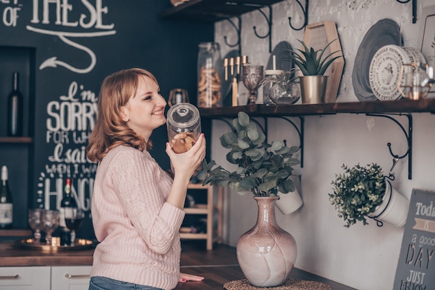 Foto de joven riendo de pie en la cocina moderna con una botella de galletas en la mano