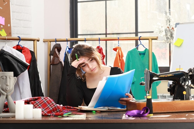 Foto de una joven sastre sentada en el taller sintiéndose cansada y mirando sus notas