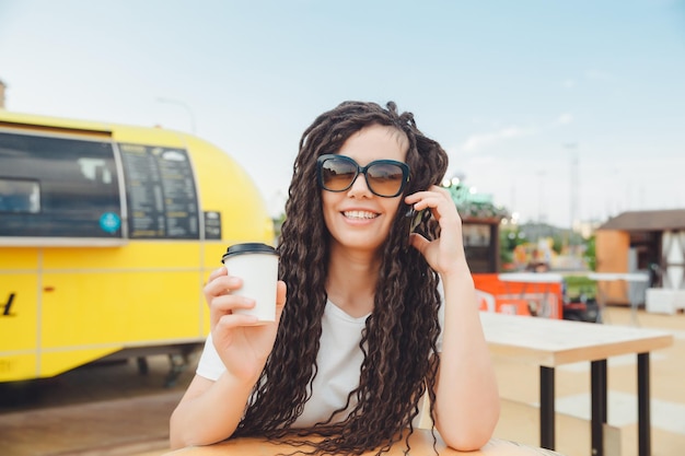 Una foto de una joven sonriente feliz que se sienta en una mesa en el patio de comidas de un centro comercial y trabaja en una computadora portátil habla en un teléfono móvil Concepto independiente