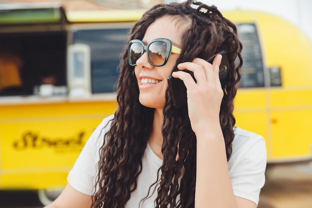 Una foto de una joven sonriente feliz que se sienta en una mesa en el patio de comidas de un centro comercial y trabaja en una computadora portátil habla en un teléfono móvil Concepto independiente