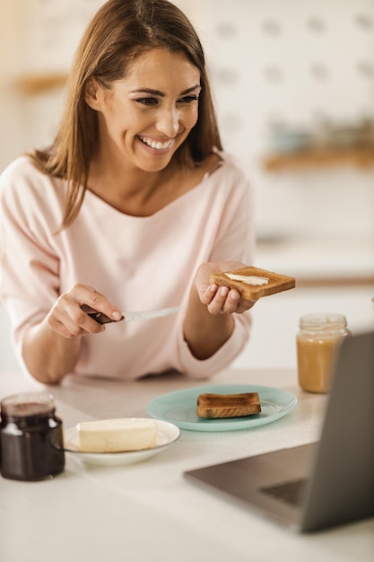 Una foto de una joven sonriente haciendo una videollamada en una laptop mientras se prepara el desayuno en su cocina.