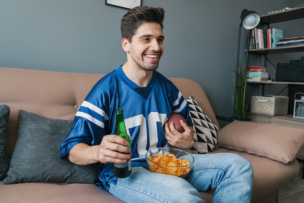 Foto de joven sonriente con pelota de rugby comiendo patatas fritas y bebiendo cerveza mientras ve el partido deportivo en la sala de estar