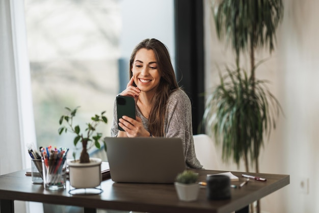 Una foto de una joven sonriente que usa un teléfono celular y una computadora portátil mientras trabaja desde casa.