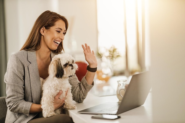 Una foto de una joven sonriente sentada con su perro mascota y haciendo una videollamada en una laptop en casa.