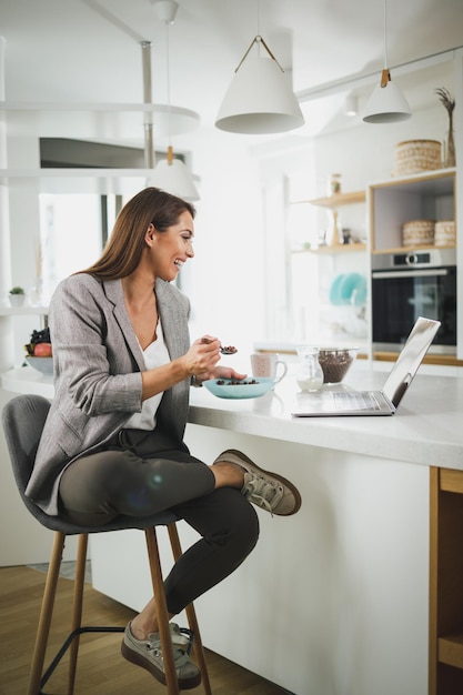 Una foto de una joven sonriente usando su laptop mientras desayunaba en su cocina mientras se preparaba para ir a trabajar.