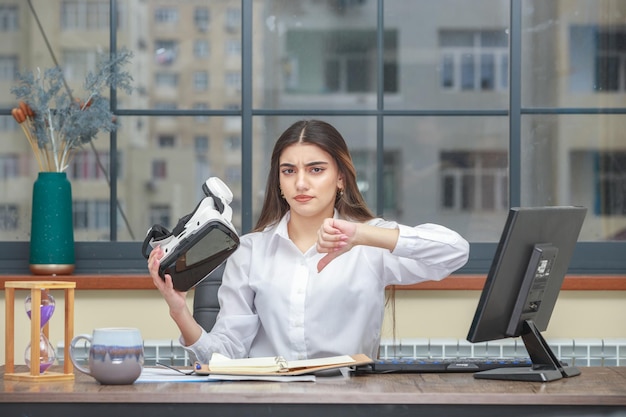 Foto de una joven sosteniendo un dispositivo de realidad virtual y haciendo un gesto con el pulgar hacia abajo