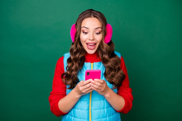 Foto de una jovencita bastante encantadora con la boca abierta sostenga el teléfono leer noticias increíbles venta de fin de semana inesperada use calentadores de oreja rosa chaleco azul jersey rojo fondo de color verde aislado