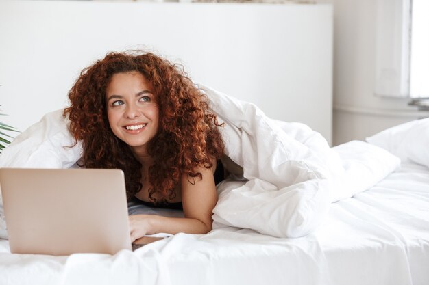 Foto de una jovencita complacida feliz pelirroja en lencería en el interior en el hotel de casa se encuentra en una cómoda cama con ordenador portátil.