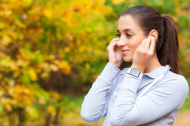 Foto de Joyful Fitness Woman 30s en ropa deportiva tocando el auricular Bluetooth y sosteniendo el teléfono móvil, mientras descansa en Green Park