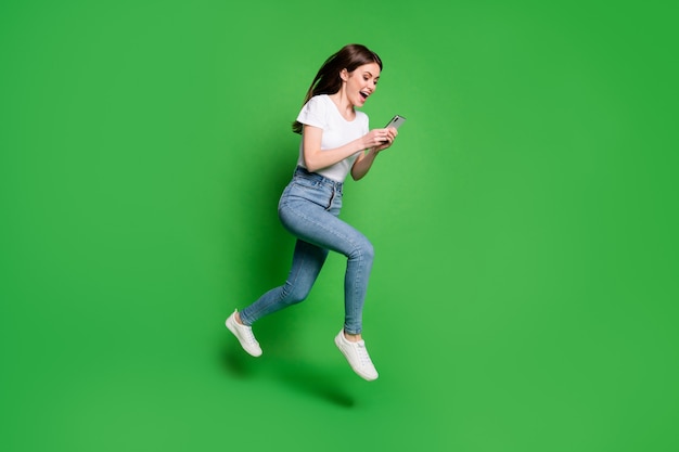 Foto lateral de perfil de corpo inteiro de garota louca, salto, corrida, leitura de smartphone usar camiseta jeans isolada de fundo de cor verde