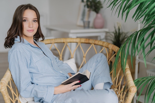 La foto lateral de una joven lectora sostiene un libro en las manos descansa en casa usa pijama disfruta de un ambiente tranquilo leyendo bestseller se sienta en una silla de mimbre Concepto de pasatiempo y relajación de la gente