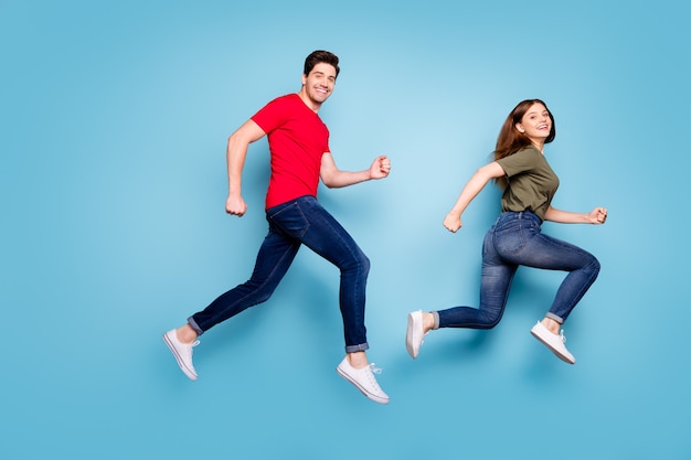 Foto lateral de perfil de tamaño completo de alegres románticos casados saltan después de los descuentos de primavera usan camiseta roja verde, jeans, zapatillas de deporte aisladas sobre fondo de color azul