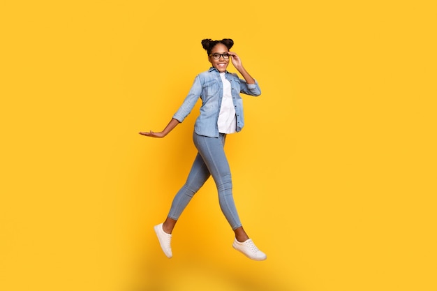 Foto lateral de perfil de tamaño completo de una joven africana que salta para caminar anteojos usa ropa informal aislada sobre un fondo de color amarillo