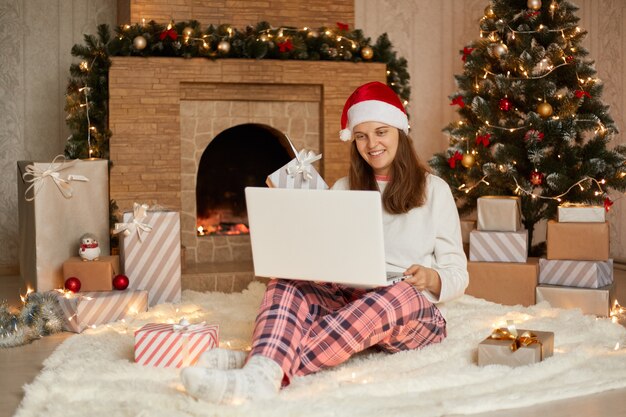 Foto de linda chica atractiva encantadora trabajando en vísperas de año nuevo con su computadora portátil o con una videollamada para saludar a alguien, vistiendo pantalones a cuadros y gorro de Papá Noel, mira la pantalla del cuaderno con una sonrisa.