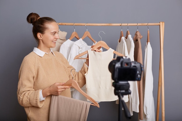 Foto de una linda estilista con suéter beige parada contra una pared gris con ropa colgada en un armario en un estante grabando video en una cámara en un trípode que muestra dos camisas diferentes