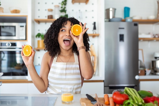 Foto de una linda mujer afroamericana sonriendo y sosteniendo dos partes naranjas mientras cocina ensalada de verduras en el interior de la cocina en casa