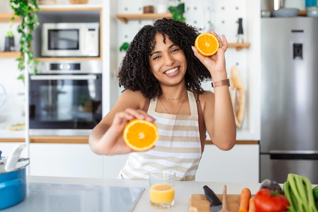 Foto de una linda mujer afroamericana sonriendo y sosteniendo dos partes naranjas mientras cocina ensalada de verduras en el interior de la cocina en casa