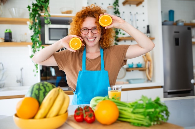 Foto de una linda mujer caucásica sonriendo y sosteniendo dos partes naranjas mientras cocina ensalada de verduras en el interior de la cocina en casa