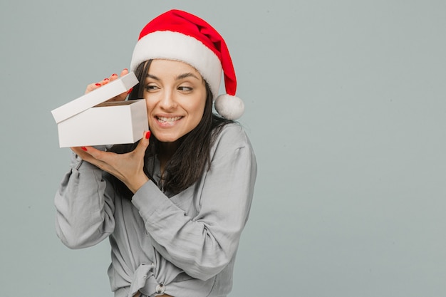 Foto foto de linda mujer sonriente con sombrero de navidad está abriendo curiosamente el presente. viste camisa gris, fondo de color gris aislado.