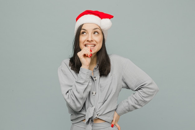 Foto de linda mujer sonriente con sombrero de Navidad parece curiosa. Viste camisa gris, fondo de color gris aislado.