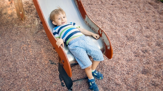 Foto de lindo niño pequeño de 3 años de edad, escalar y montar en tobogán grande en el parque infantil en el parque