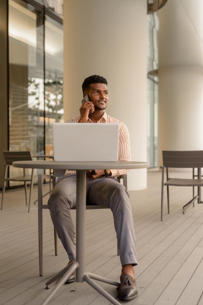 Foto de longitud completa del empresario africano en la ciudad con ordenador portátil en la cafetería mientras habla por teléfono