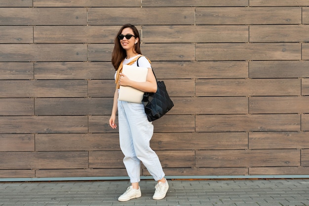 Foto de longitud completa de la joven y encantadora mujer morena hermosa que va en la calle cerca de la pared marrón