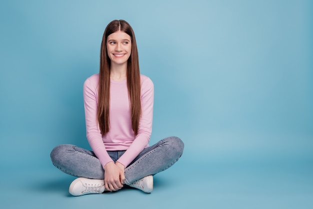 Foto de longitud completa de la niña feliz sonrisa positiva sentada en el suelo sueño mirar espacio vacío aislado sobre fondo azul.