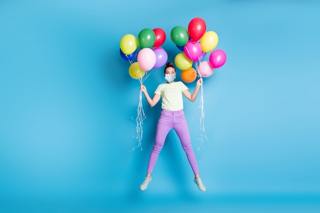 Foto de longitud completa de niña sorprendida saltando alto con globos mirando máscara de desgaste aislada sobre fondo de color azul brillante