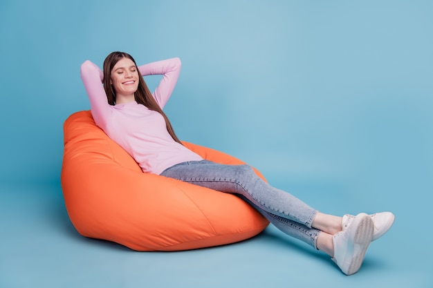 Foto de longitud completa de la relajante niña sentada en el sillón puf sueño mano soñadora detrás de la cabeza aislada sobre fondo de color azul