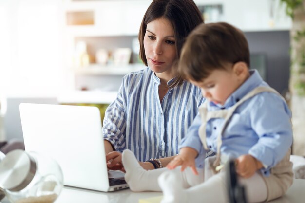 Foto de madre bastante joven que trabaja con la computadora portátil mientras su bebé está sentado en la encimera de la cocina en casa.