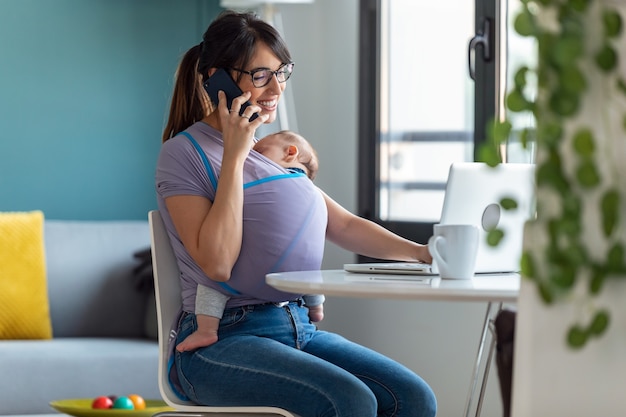 Foto de madre bastante joven con su bebé en cabestrillo hablando por teléfono móvil mientras trabaja con la computadora portátil en casa.