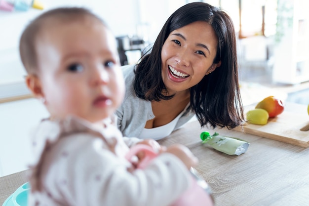 Foto de madre joven feliz con su hija mirando a cámara en la cocina de casa.