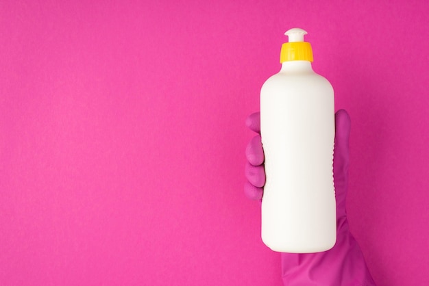 Foto de mano en guante de goma rosa sosteniendo una botella de espuma de detergente sin etiqueta sobre fondo rosa aislado con espacio de copia