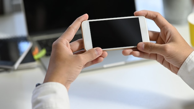 Foto de la mano del joven empresario sosteniendo un teléfono inteligente blanco con pantalla en blanco negro en la mesa de trabajo moderna con borrosa portátil y equipo de oficina como