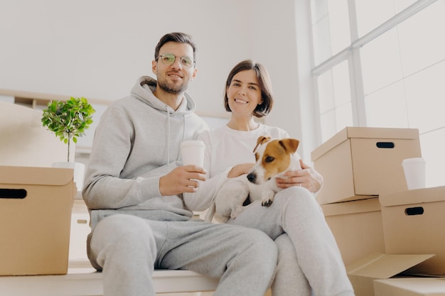 Una foto de un marido y una mujer alegres posan junto con un perro de pedigrí en una habitación vacía sin muebles, cajas de cartón con pertenencias personales, beben café para llevar, disfrutan de la unión